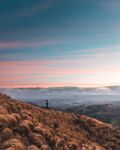 Breathtaking landscape capturing a woman on a mountain, enjoying clean air as she gazes down at a valley where clouds hover. The sunset paints the clouds in hues of pink and orange, creating a tranquil scene.