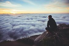 A serene moment captured as a man sits on the summit of a mountain, gazing down at the valley blanketed in clouds, with the sun setting on the horizon, casting a warm glow.