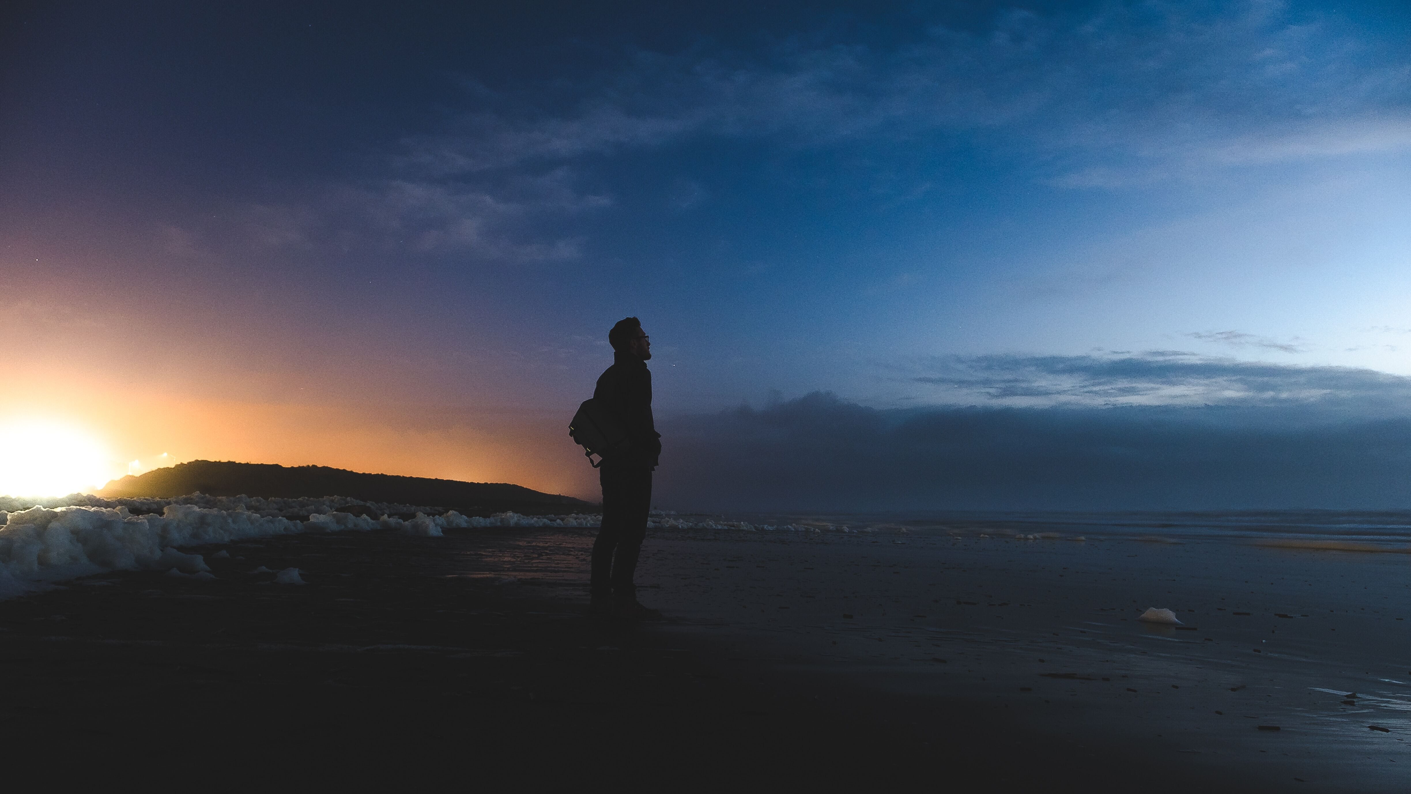 A serene moment captured on the beach as a man stands, looking out to the sea, with the sun setting behind a mountain, casting a warm glow.