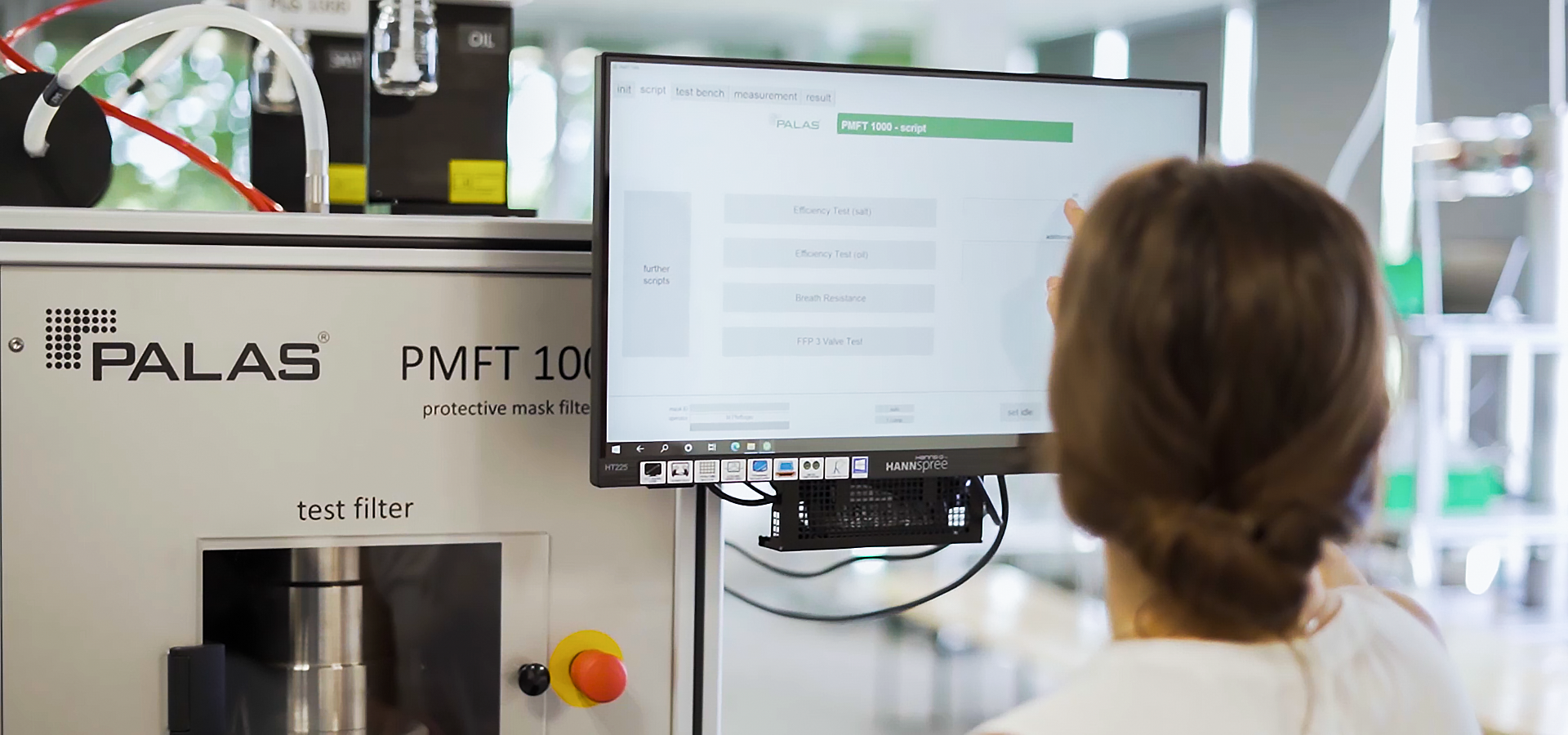 A female researcher wearing a white coat in a laboratory works on a monitor of the Palas&reg; device; the PMFT 1000 allows the development and production monitoring of face masks.