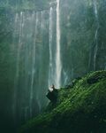 A serene scene capturing a man seated on a rock ledge, looking up towards a lush green forest, while behind him, a towering rock wall adorned with cascading waterfalls adds to the breathtaking natural setting.