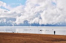 Idyllic beach scene featuring a woman enjoying the pristine air quality as she gazes across a serene lake with boats, ducks, and rocks. Majestic mountains stand tall in the background, and low-hanging clouds touch the still water, creating a picturesque s