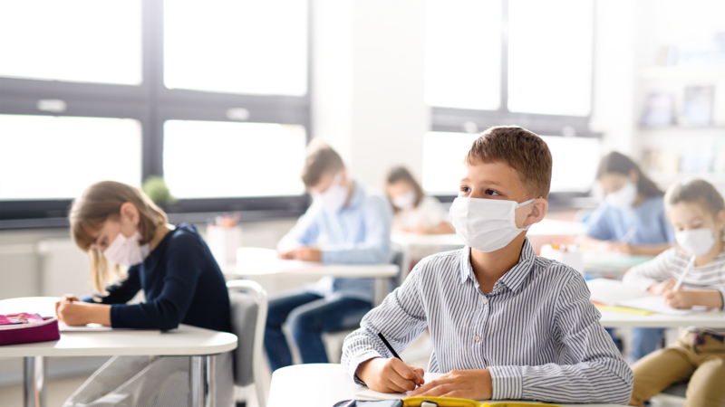 Six children wearing masks sit at their school desks, the young boy in the front looks to the front. Palas offers measuring devices to monitor the indoor air quality, in schools, work spaces or laboratories.