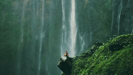 Inhaling the freshness of the great outdoors, a man sits on a rock ledge, surrounded by the calming sights and sounds of a vibrant green forest and cascading waterfalls against a massive rock wall.