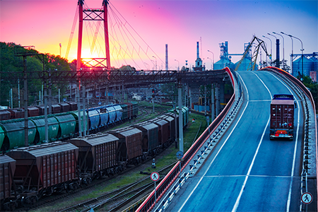 A picturesque scene capturing a truck crossing a bridge against the backdrop of a sunset. On the left, a railway and industrial buildings provide a dynamic context to the industrial setting.
