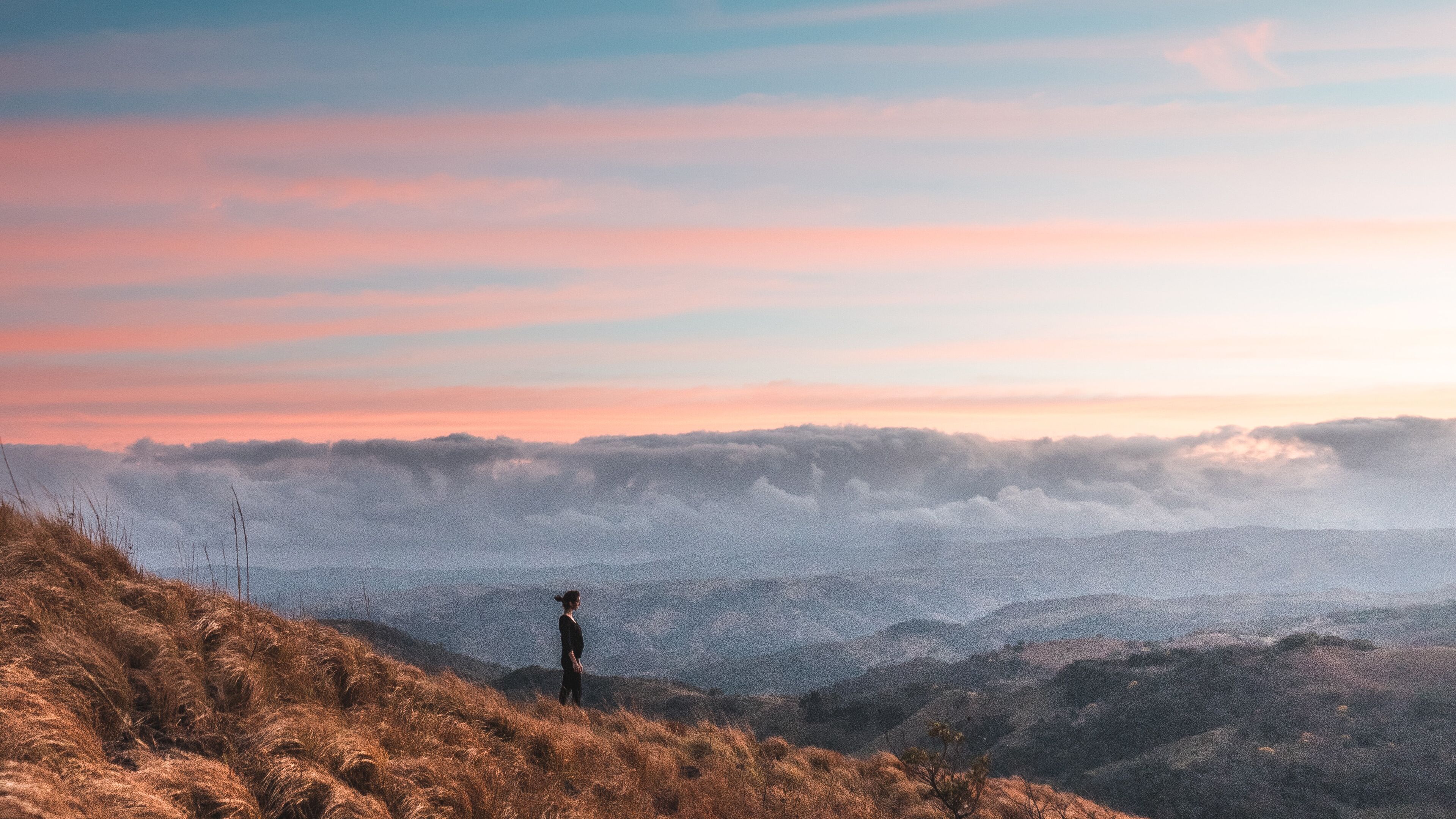 A woman's profile as she stands on a mountain summit, breathing in the crisp, clean air, with the valley below blanketed in clouds painted in the warm hues of a sunset sky.